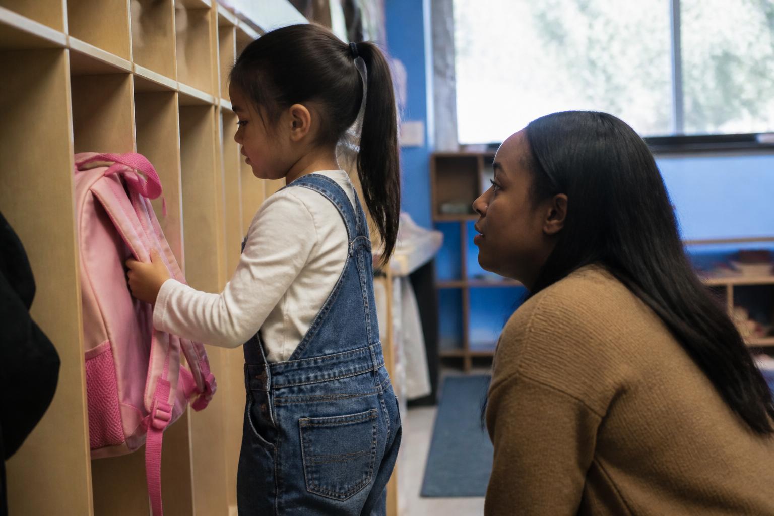 A Friend kneels at eye level beside a young child hanging a backpack in a Houston classroom cubby.